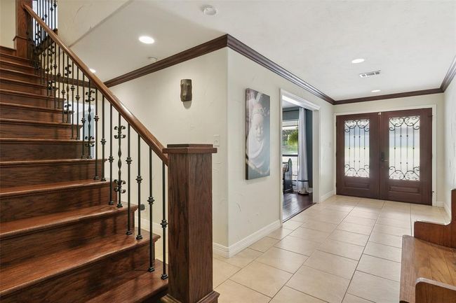 The photo showcases a spacious, inviting foyer with elegant wooden stairs and a wrought-iron railing. The area features tiled flooring, decorative wall molding, and stylish double doors with glass inserts, creating a warm and sophisticated entrance. | Image 36