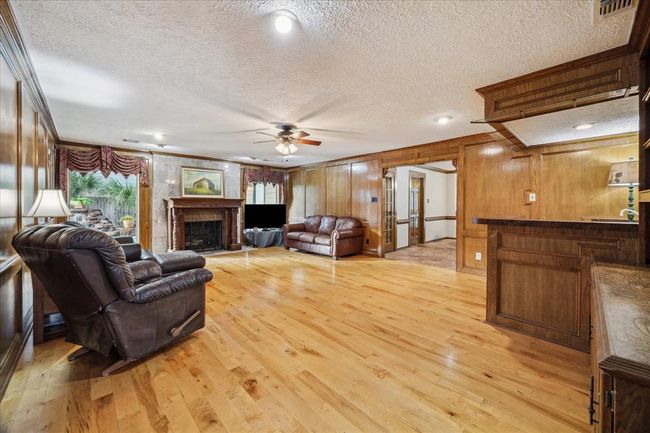 This spacious living room features warm wood paneling and maple flooring, a cozy marble fireplace, and ample natural light from large windows. It includes a wet bar area, making it ideal for entertaining. | Image 7
