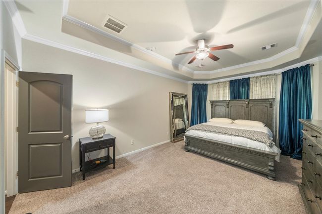 The primary bedroom boasts a tray ceiling with a fan, soft neutral carpeting, and a large window adorned with blue curtains. | Image 17