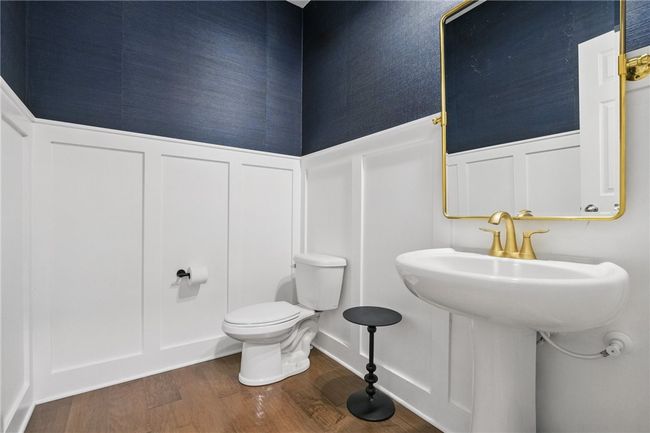 Stylish half bath featuring elegant two-tone walls with white wainscoting and deep navy above. Includes a pedestal sink with brushed gold fixtures and matching mirror, all set atop warm-toned hardwood flooring for a refined, modern look. | Image 7