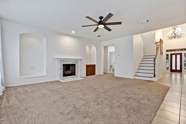 Spacious living room with a cozy fireplace, neutral carpeting, and a modern ceiling fan. It features arched wall niches, a stairway leading upstairs, and an open layout that flows into a tiled entryway with a chandelier. | Image 9