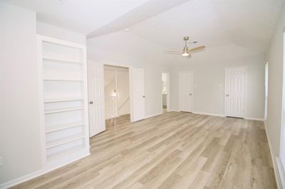 This is a bright, spacious primary bedroom with modern light wood-look flooring, fresh white paint, and a built-in shelving unit to the left. The room features a ceiling fan and high ceilings. | Image 3