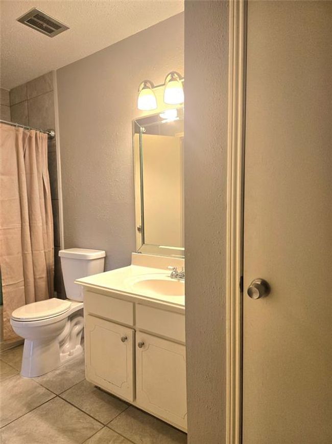 This bathroom features a simple design with a white vanity, mirror, and overhead lighting. It includes a toilet and a shower with a curtain, all set against neutral-toned walls and tiled flooring. | Image 11
