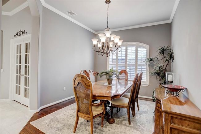 An elegant dining room features a chandelier and large windows with plantation shutters, set on a neutral color palette with crown molding. | Image 7