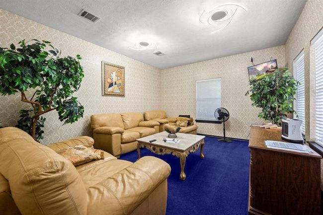 This is a well-lit, cozy living space featuring a matching tan sofa set and a classic coffee table. The room has a vibrant purple carpet, textured wallpaper, and is accented with indoor plants and wall art. The space is equipped with two windows for natural light and a ceiling fan for comfort. | Image 29