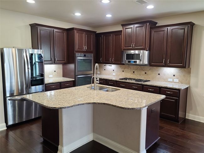 Kitchen with stainless steel appliances, dark wood-style floors, dark brown cabinets, light stone counters, and a center island with sink | Image 13