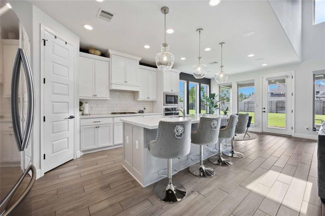This modern kitchen features sleek white cabinetry, a large island with stylish barstools, and elegant pendant lighting. Ample natural light streams in through large windows and a glass door, providing views of a lush backyard. | Image 13