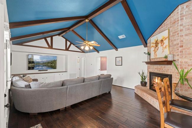 This inviting living room features a high vaulted ceiling with exposed beams, a striking blue color, and includes a cozy brick fireplace. The space is accented with dark hardwood floors and an overhead ceiling fan, creating a warm and stylish atmosphere. | Image 13