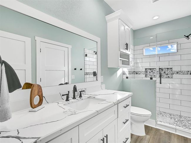 Relaxtion awaits in this elegant en suite master bath featuring a vanity with quartz countertops, a rectangular basin sink, custom cabinetry, a framed mirror, and a spacious walk-in shower with decorative tile accents and frameless glass. | Image 19