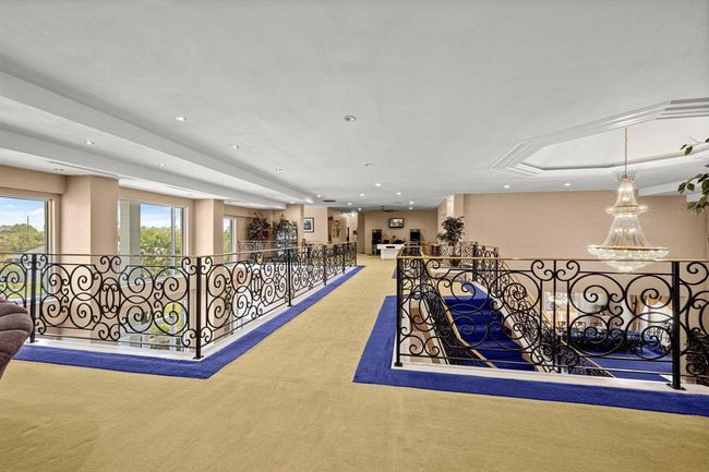 This is a spacious, well-lit interior space featuring an elegant open floor plan with a grand staircase, ornate metal railings, and large windows that provide ample natural light and views of the surrounding area. A classic chandelier adds a touch of luxury to the room. | Image 20