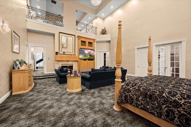 This is a spacious, elegantly appointed primary bedroom with high ceilings, large windows, and an open second-level balcony. It features a comfortable seating area, a fireplace, and patterned carpeting, creating a luxurious and inviting atmosphere. | Image 13