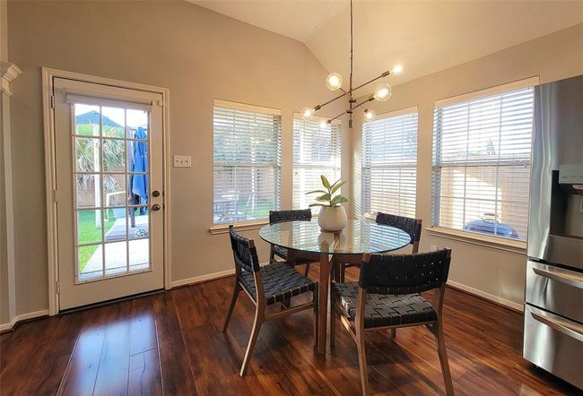 Bright dining area with hardwood floors, featuring a modern chandelier above a round glass table with four chairs. Large windows and a glass door provide ample natural light and a view of the backyard. | Image 11