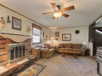 Living room with a brick fireplace, a textured ceiling, light colored carpet, ceiling fan, and wooden walls | Image 3