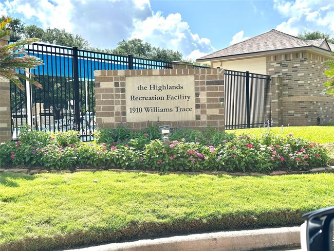 This photo shows the entrance to The Highlands Recreation Facility at 1910 Williams Trace. The facility is surrounded by a well-maintained lawn and flower beds, with a brick sign and a secure black metal fence. The structure in the background suggests additional amenities. | Image 12