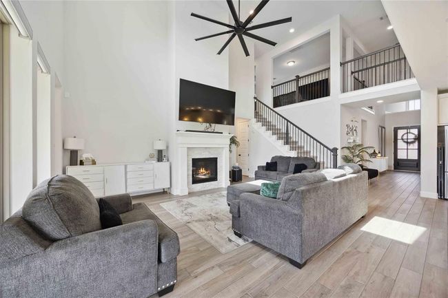 This spacious living room features a high ceiling with a large ceiling fan, a cozy fireplace, modern furnishings, and an open staircase leading to the upper level. Natural light fills the room through large windows, creating a bright and inviting atmosphere. | Image 11