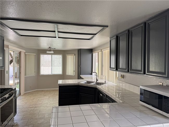 Kitchen with tile counters, white microwave, stainless steel range with electric stovetop, dark cabinetry, and a textured ceiling | Image 6