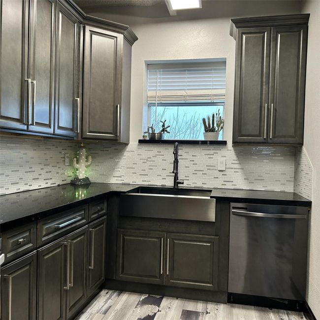 This kitchen features sleek dark cabinetry, a modern farmhouse sink, and stainless steel appliances. It includes a stylish tile backsplash and a window above the sink, providing natural light and a view. The design combines functionality with contemporary aesthetics. | Image 14