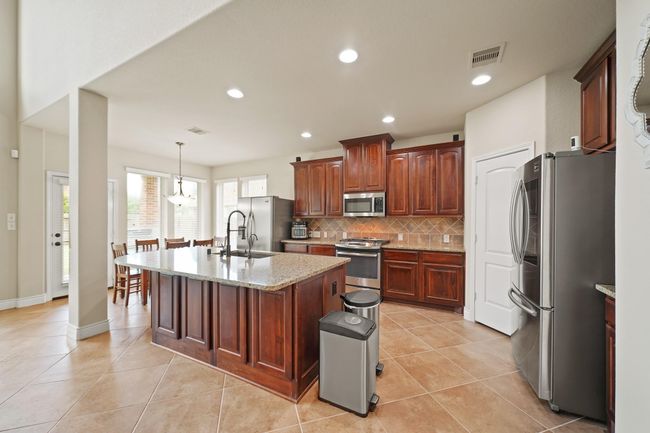 This spacious kitchen features elegant wooden cabinetry, a large central island with granite countertops, stainless steel appliances, and tile flooring. The open layout flows into a bright dining area with natural light from large windows and access to the patio. (Refrigerators do not convey). | Image 15