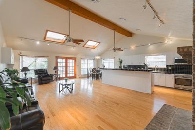 This is a spacious, open-concept living area with vaulted ceilings, abundant natural light, hardwood floors, and large kitchen with white cabinetry and black countertops. The space is well-lit with a combination of track lighting and skylights. | Image 1