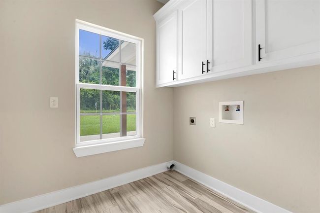 This is a bright laundry room featuring large windows, white cabinets with black handles, and hookups for a washer and dryer. The room has light-colored walls and flooring, creating a clean and inviting space with a view of greenery outside. | Image 17