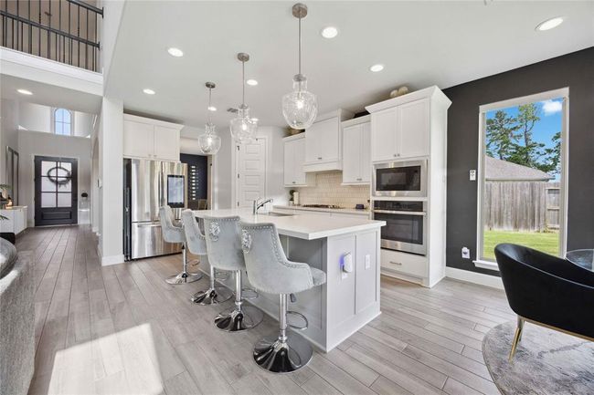 This modern kitchen features a spacious open layout with a large island, elegant pendant lighting, and sleek stainless steel appliances. White cabinetry and light wood flooring create a bright, inviting atmosphere, complemented by a view of the backyard through a large window. | Image 14