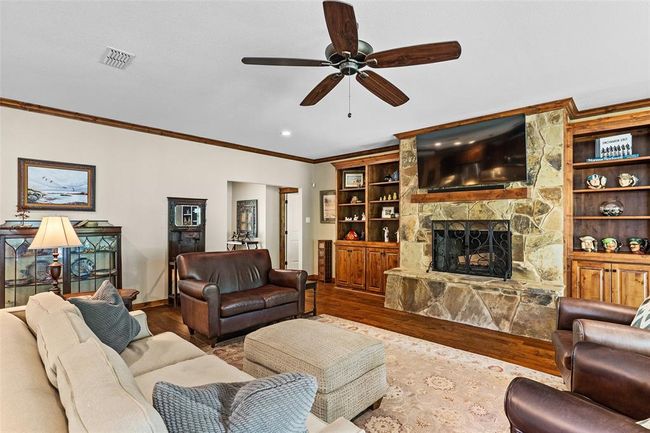 Living room with wood-type flooring, crown molding, built in shelves, a stone fireplace, and ceiling fan | Image 4