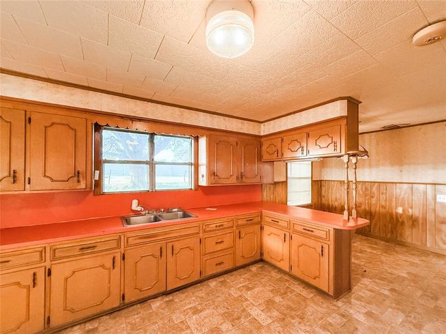 This photo shows a vintage kitchen with wood cabinets and bold red countertops. It features a double sink under a window, ample storage, and a cozy, retro feel with wood paneling and patterned flooring. | Image 7