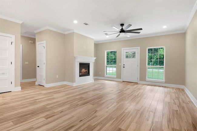 This spacious living room features elegant wood flooring, a cozy white brick fireplace, and ample natural light from large windows. A modern ceiling fan adds comfort, and the neutral walls create a warm, inviting atmosphere. | Image 12