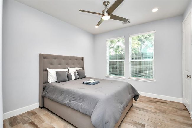 This bedroom features a neutral color palette with light gray walls and wood-look tile flooring. It includes a comfortable double bed with a tufted headboard, complemented by modern decor. Large windows provide ample natural light, and a ceiling fan adds comfort. | Image 11