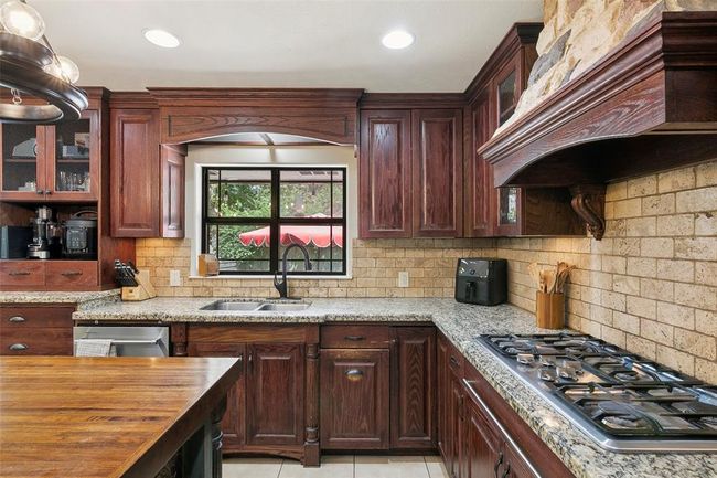 This kitchen features rich wooden cabinetry, granite countertops, and a built-in gas stove. It includes a central island and a window with a view, providing ample natural light. The backsplash is made of stone tiles, adding a rustic touch. | Image 21