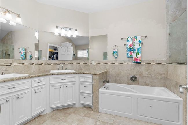 This is a spacious bathroom featuring a large mirror, double vanity with white cabinetry, a built-in bathtub, and tiled walls. It has a neutral color palette and good lighting. | Image 29