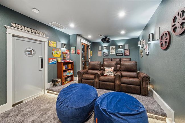 This dedicated movie room is designed for the ultimate cinematic experience. The movie-style swinging door with a window and a cinema marquee outside sets the stage for what’s inside. Inside, plush carpet underfoot, custom lighting (including a built-in on-lighting system), and two elevated seating platforms ensure the perfect view for everyone. A glass-doored mechanical closet keeps everything organized, while the projector TV and surround sound system create the perfect environment for movie nights. | Image 28