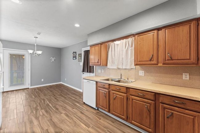 This photo shows a spacious kitchen and dining area with wooden cabinetry, and neutral walls. It features a double sink, dishwasher, and a dining nook with a chandelier. A door leads to an outdoor space, providing ample natural light. | Image 18