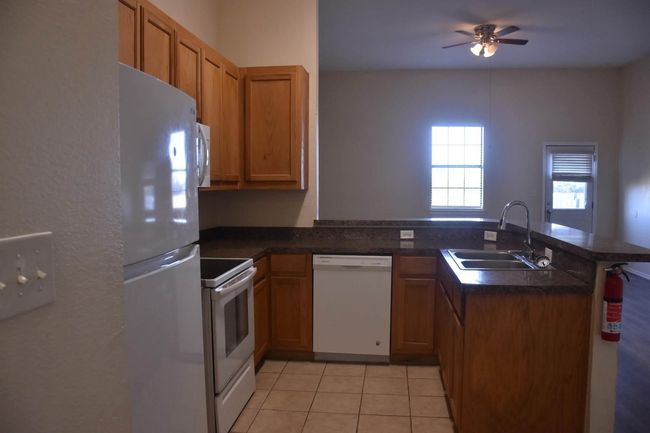 Kitchen with dark countertops, a peninsula, white appliances, a ceiling fan, and a sink | Image 7