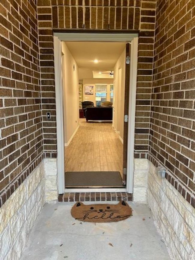 This photo shows a welcoming entryway with a brick exterior and a "hello" doormat. The open door leads to a well-lit interior with wood-like tile flooring, offering a glimpse into a cozy living area. | Image 4