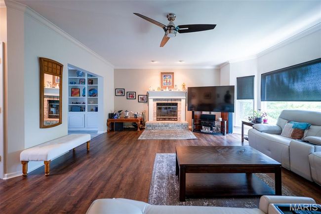 Living room with a fireplace, crown molding, a ceiling fan, wood finished floors, and built in shelves | Image 13