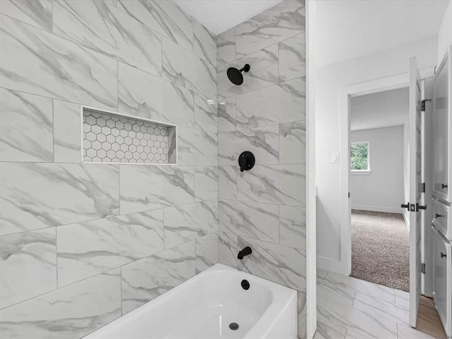This bathroom features a modern bathtub with elegant marble-style wall tiles and matte black fixtures. It includes a hexagonal tile niche for storage and an adjoining bedroom view. | Image 32