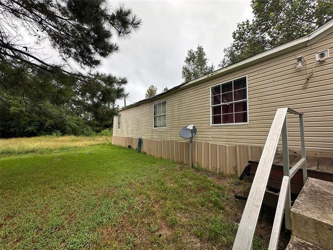 This photo shows a single-story mobile home with light siding, situated on a grassy lot surrounded by trees. A satellite dish is mounted outside, and there are steps leading to an entrance. The setting provides a sense of privacy and nature. | Image 10