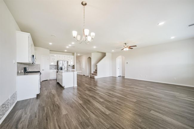This spacious open-concept living area features sleek wood flooring, a modern kitchen with white cabinetry, stainless steel appliances, and an island. The room is bright and airy, with recessed lighting, a ceiling fan, and a stylish chandelier. A staircase leads to the upper level, enhancing the home's elegant design. | Image 16