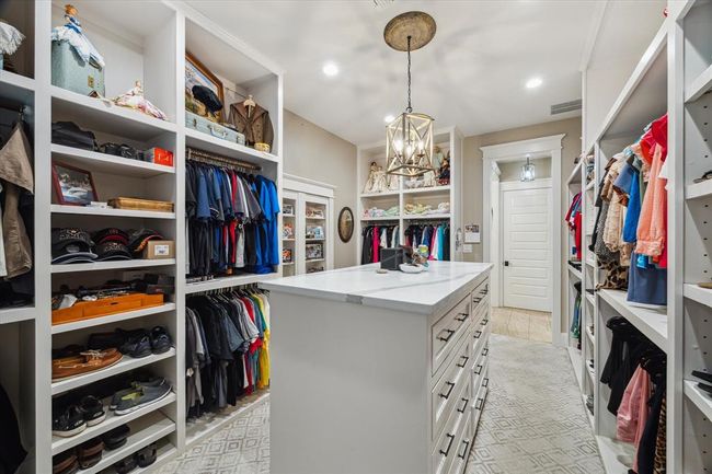 The primary closet offers a refined touch with custom high-end designer carpeting, built-in shelving, and a built-in dresser with sleek quartz countertops. A stylish sitting bench provides a convenient space to relax or get ready, while an elegant chandelier casts a warm glow, completing the luxurious feel of this well-appointed closet. | Image 20