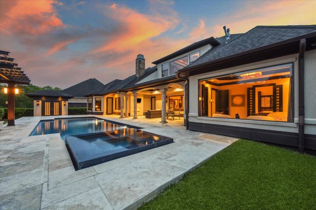 Surrounded by glowing windows and stately stone columns, this twilight view captures the seamless flow between the luxurious indoor living spaces and the serene backyard oasis. The elevated spa spills gently into the sparkling pool, while the surrounding travertine decking enhances the elegant aesthetic. Soft uplighting from the pergola and fire pit lounge creates a magical ambiance, perfect for evening entertaining or quiet relaxation under the stars. | Image 43