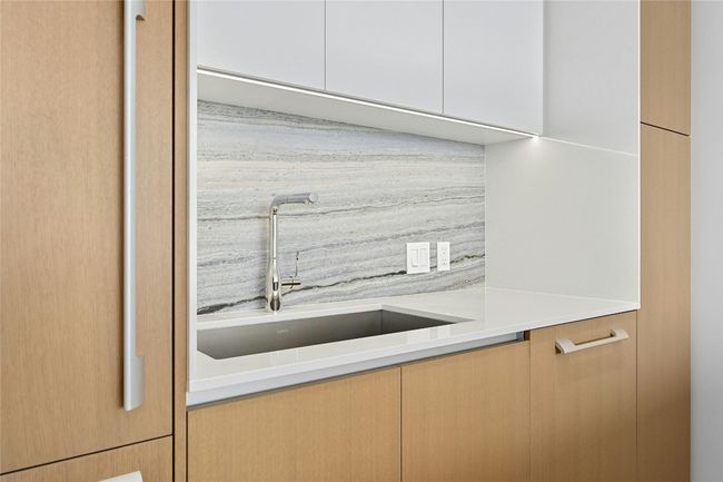 The kitchen features a sleek, minimalist design with clean-lines cabinetry that combines light wood finishes and white fronts. The backsplash matches the island countertop, creating a cohesive and sophisticated look. | Image 5
