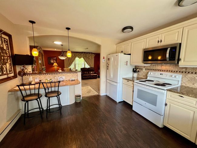 Kitchen with white appliances, a baseboard radiator, dark wood finished floors, a kitchen bar, and white cabinets | Image 6