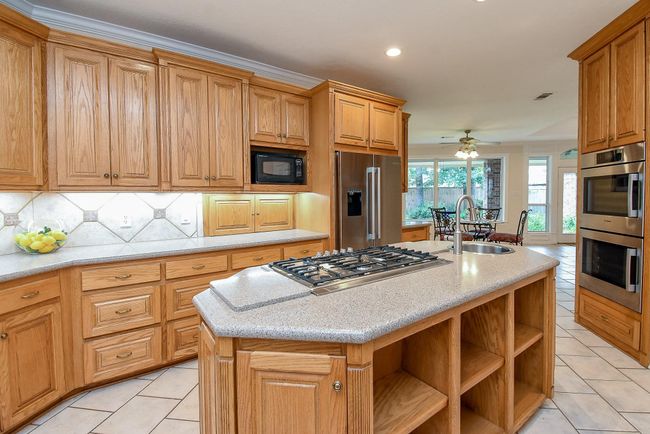 Spacious kitchen with ample wooden cabinetry, modern stainless steel appliances, and a central island featuring a gas cooktop and sink. The open layout includes a dining area with large windows for natural light. | Image 18