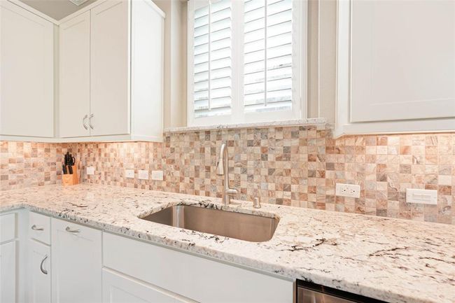 Close-up of kitchen showcasing granite countertops, under-cabinet lighting, stainless steel undermount sink, and mosaic tile backsplash. A blend of function and style with ample cabinetry and natural light. | Image 15