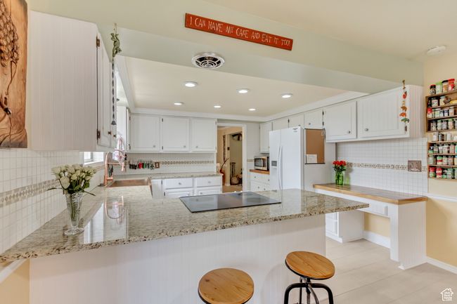 Kitchen featuring a peninsula, stainless steel microwave, backsplash, white refrigerator with ice dispenser, and cooktop | Image 13
