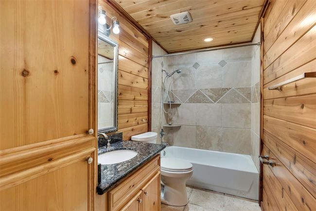 This bathroom features a cozy rustic design with wood-paneled walls and ceiling. It includes a granite countertop with a sink, a mirror with overhead lighting, and a tiled shower-tub combination. | Image 46