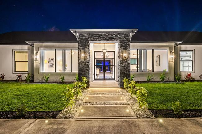 This stunning single-story residence showcases sleek architectural lines, expansive picture windows, and a grand stone-accented entryway illuminated by warm, ambient lighting. The manicured landscaping and illuminated walkway create an inviting path to the glass-paneled front door, offering a glimpse of the sophisticated interiors beyond. Designed for luxurious living and effortless entertaining, this home blends contemporary style with timeless comfort. | Image 6