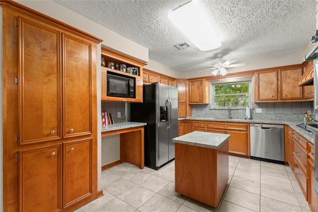 This kitchen features warm wood cabinetry, a small central island, and a double oven set in a brick accent wall. It has stainless steel appliances, granite countertops, and a window above the sink. The open layout connects to a cozy living space. The island is mobile and can be rolled back into the cabinet. | Image 10