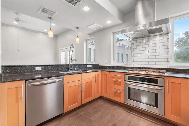 Kitchen featuring stainless steel appliances, wall chimney range hood, wood finished floors, tasteful backsplash, and dark stone countertops | Image 5